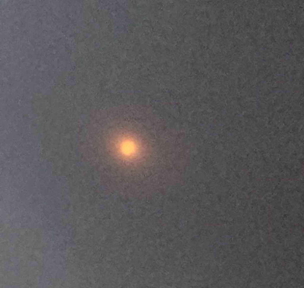 First bite image of eclipse taken through flimsy "eclipse glasses". The image is mostly black with a small 'bite' mark out of a red sun's lower right side.