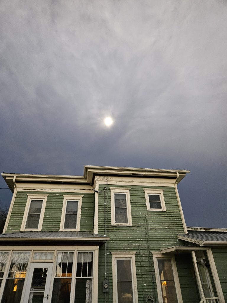 Photo of totality approaching as a dark band coming from the west. It almost appears as though the clouds have formed a hand that is reaching out to grab the sun. The sky is distinctly dark as the moon's shadow rapidly approaches, even though there is a lot of cloud cover.